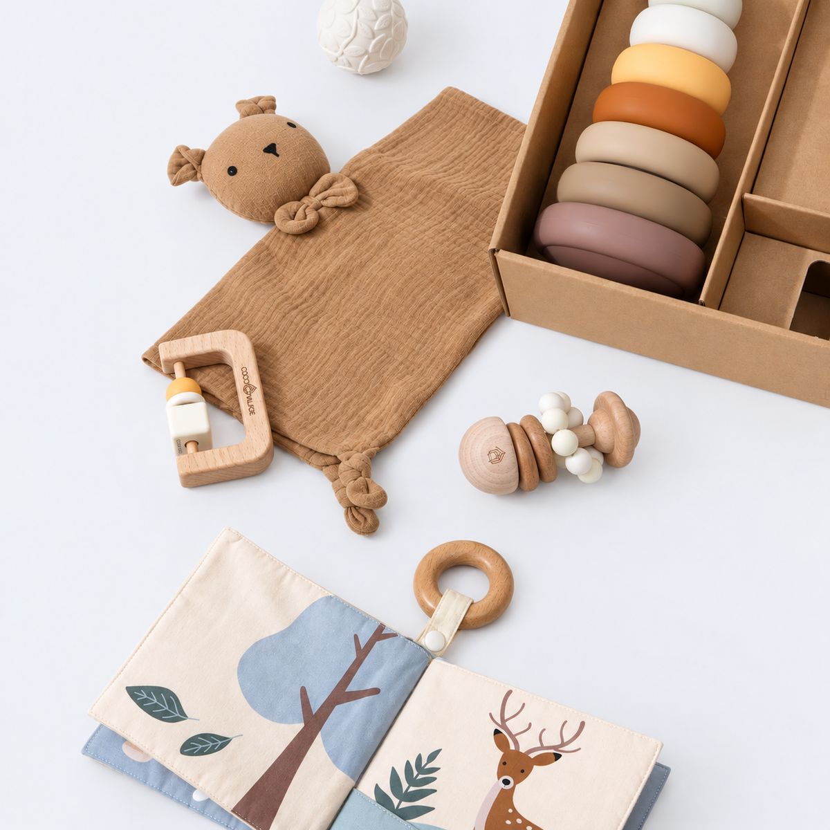 A baby gift set arranged on a white background, including a brown muslin swaddle with a plush bear, a wooden teething ring, a wooden bottle holder, a wooden teething toy, a box of stacked wooden rings, and a fabric baby book with tree and deer illustrations.