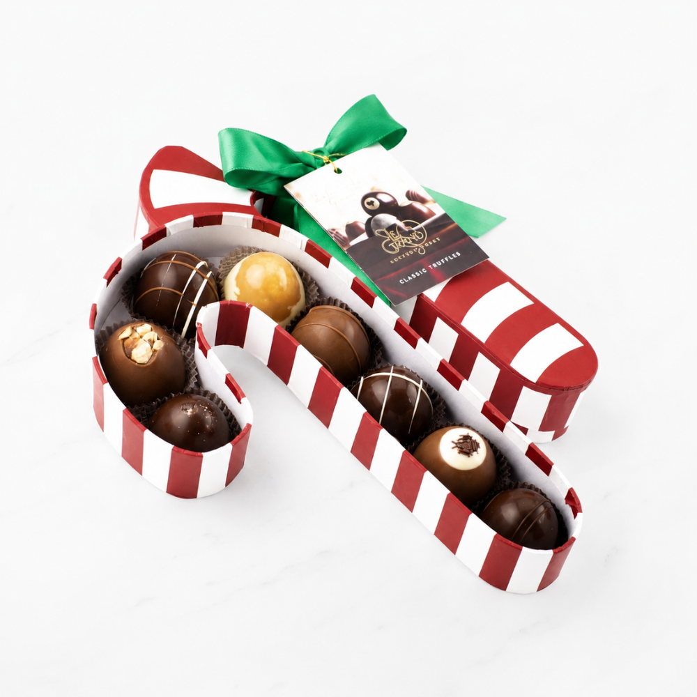 A box of assorted chocolates arranged in a red and white striped candy cane shape, tied with a green ribbon and a chocolate tag, on a white marble background.