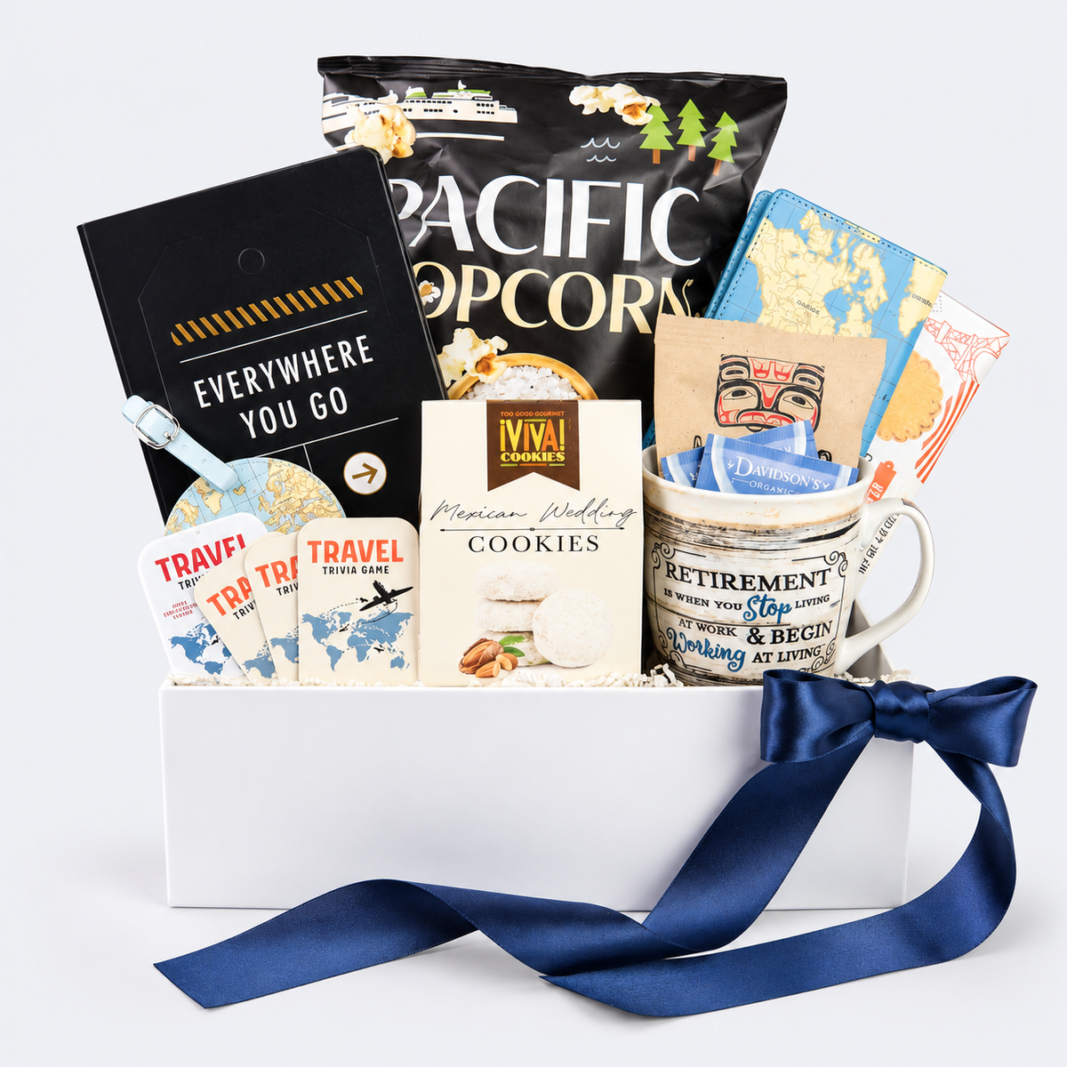 A curated gift basket filled with travel-themed items, including a bag of Pacific Popcorn, Mexican Wedding cookies, travel cards, a mug with a 'Retirement' design, and a black 'Everywhere You Go' card, arranged in a white box with a navy blue ribbon on a light gray background.