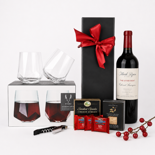 A curated gift set on a white studio background featuring a bottle of Mark Opes Red Label Winery wine, two faceted clear wine glasses in and out of their box, a tall black gift box tied with a red bow, smoked applewood cigar spruce boxes, red tea bags, a corkscrew, and red berries.