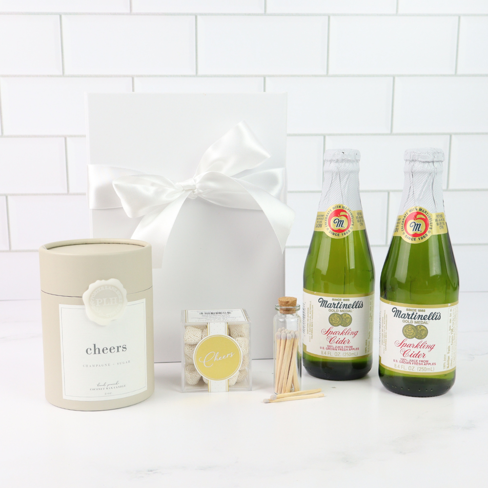 Two bottles of Martinellis sparkling cider with a candle labeled 'cheers' and a gift bag on a white tiled background.