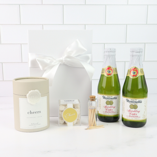 Two bottles of Martinellis sparkling cider with a candle labeled 'cheers' and a gift bag on a white tiled background.