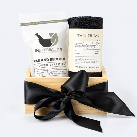 A gift set in a light wood box tied with a black ribbon, containing a white bag of The Herbal Zen Shower Steamers and a black rolled Tea With Tae towel.